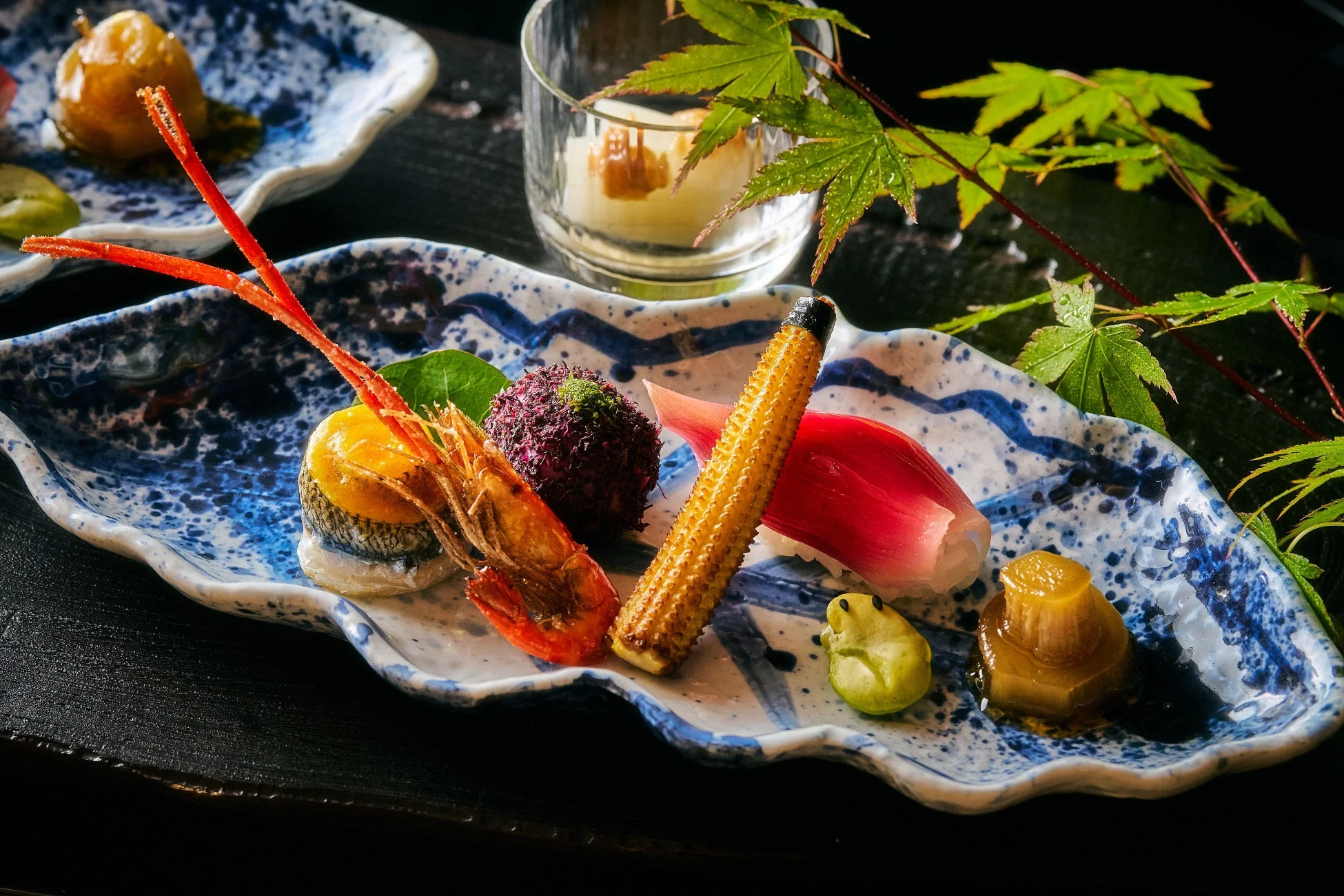 青と白の美しい器に盛り付けられた、海老やヤングコーンなど色とりどりの和食前菜。水滴のついた青もみじが添えられ、季節感あふれる上品な会席料理の一品を示しています。