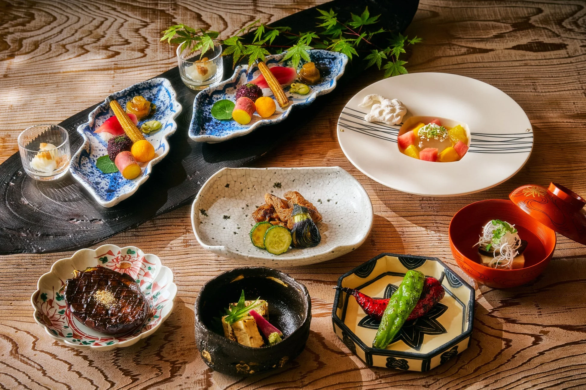木製のテーブルに美しく盛り付けられた和食のコース料理が並んでいます。色とりどりの前菜、焼き茄子、スープ、フルーツゼリーなどが、もみじの葉と共に上品に配置されています。