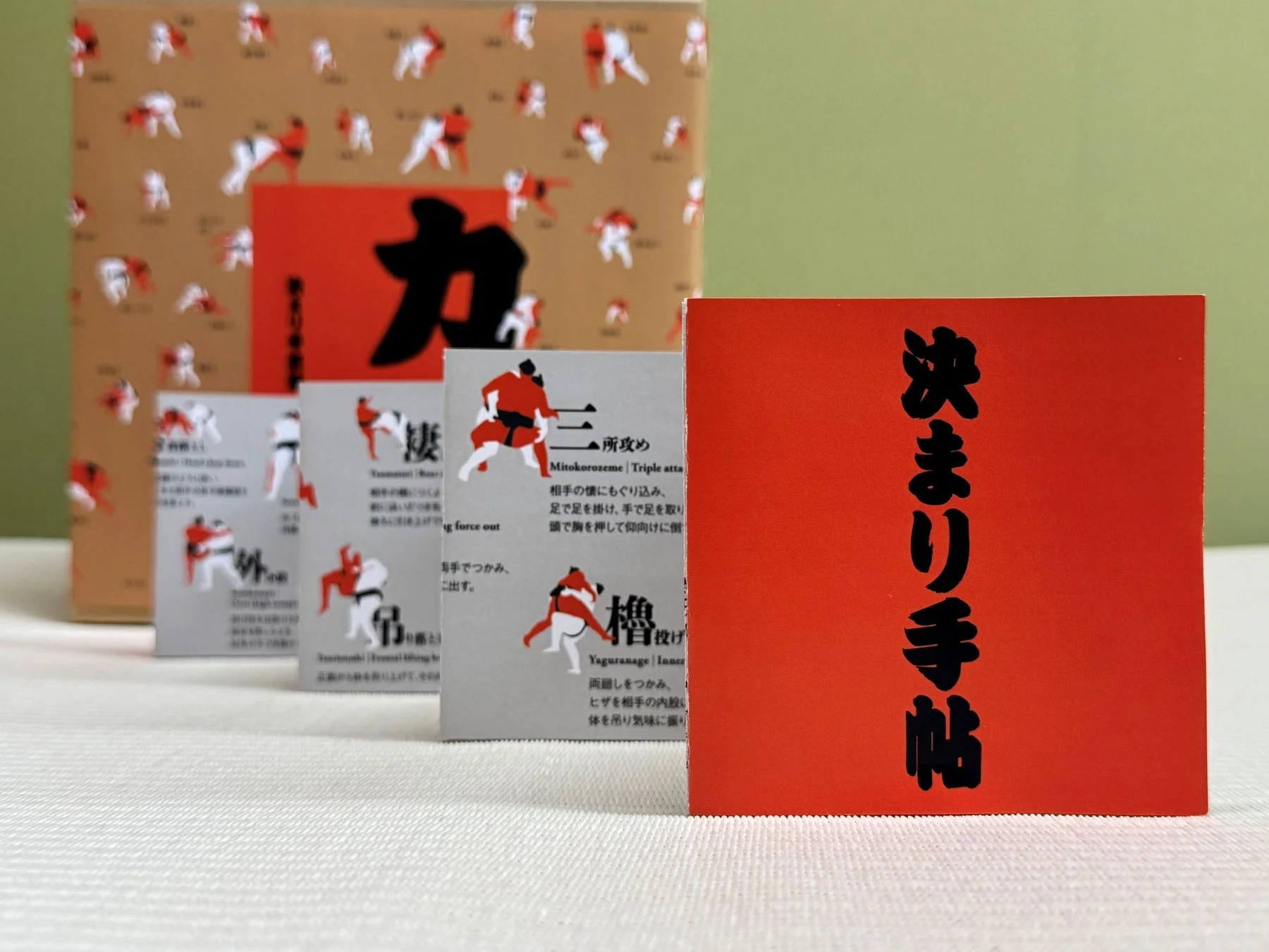 柔道や相撲の「決まり手」を解説した冊子やパッケージが並べられています。赤い表紙には「決まり手帖」と書かれ、技のイラストと共に詳細な説明が記されています。