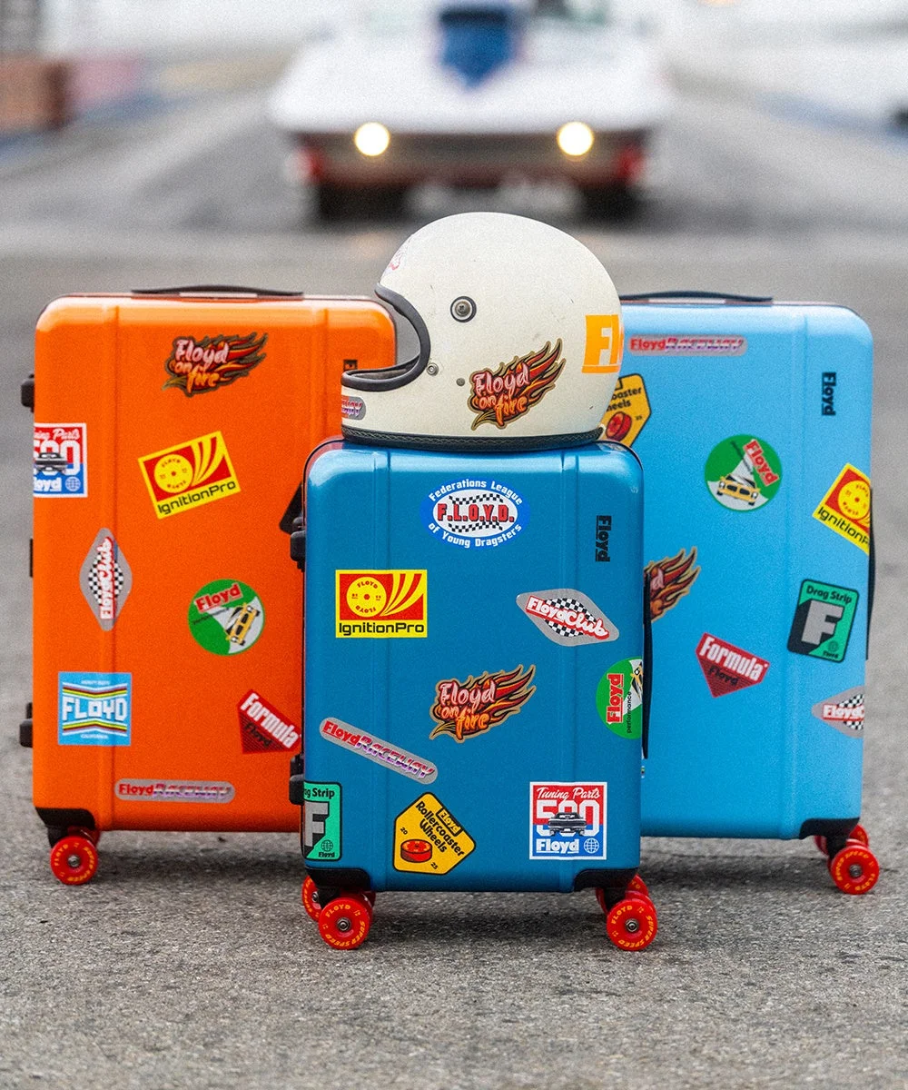 レトロなレーシングステッカーで飾られたカラフルなスーツケース3個とヘルメットが並べられ、背景にはクラシックカーが見える