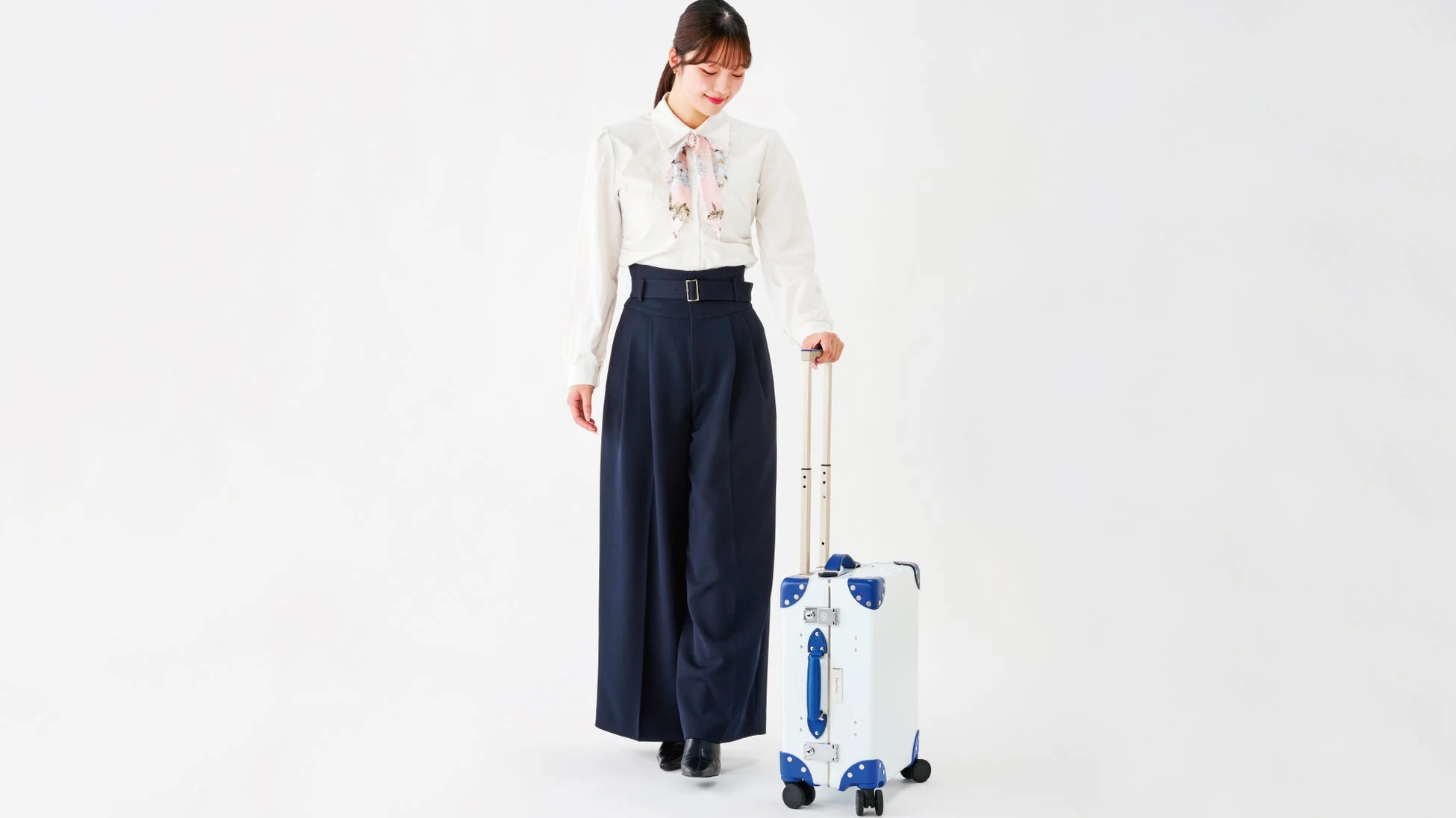 白いシャツとネイビーのワイドパンツを着用した女性が、白いスーツケースを持って立っています。