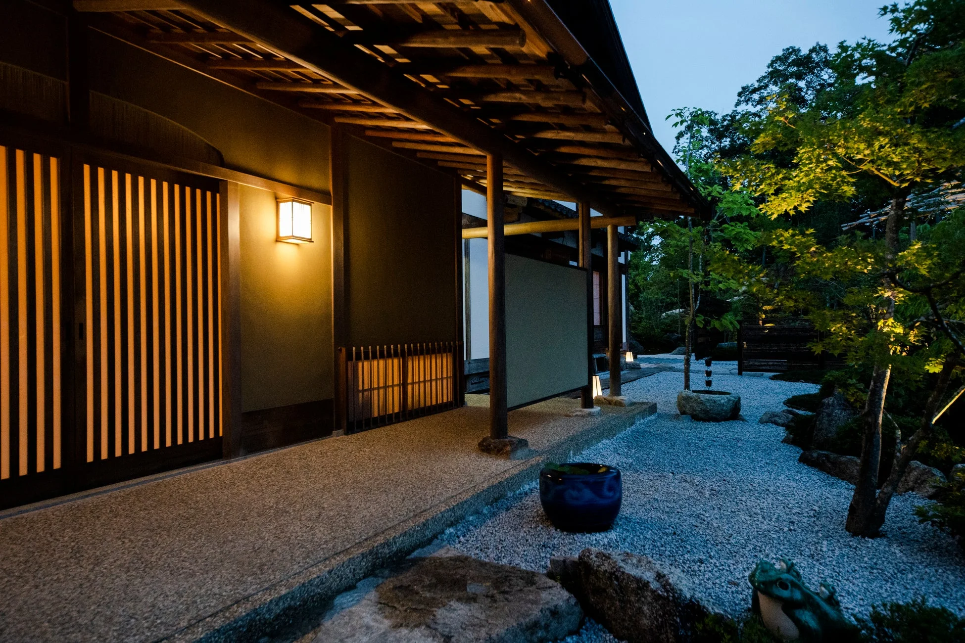 夕暮れ時か夜の日本庭園と和風建築の様子。温かい光を放つ門灯が玄関を照らし、石灯籠や庭園灯でライトアップされた砂利敷きの庭が幻想的な雰囲気を醸し出しています。