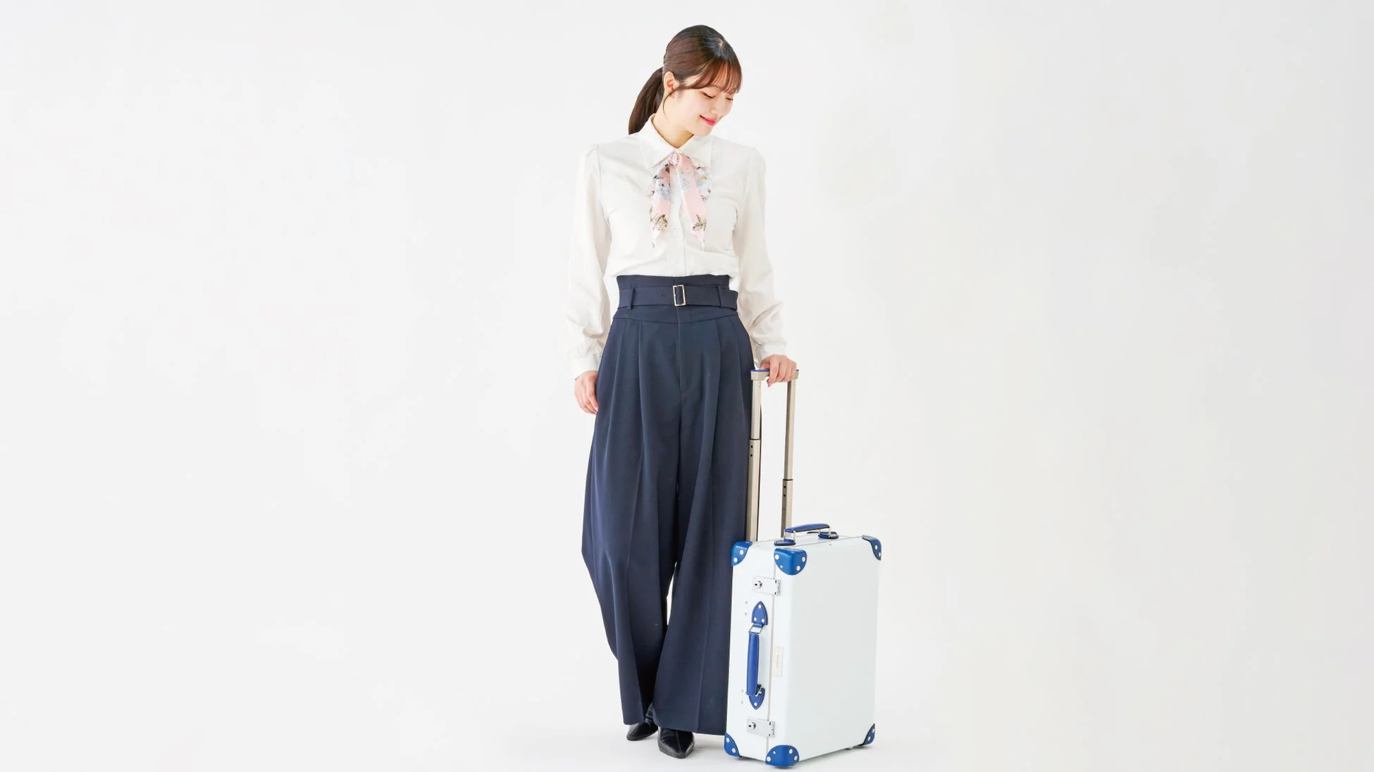 女性が白いシャツとネイビーのワイドパンツを着用し、白いスーツケースを持って立っています。